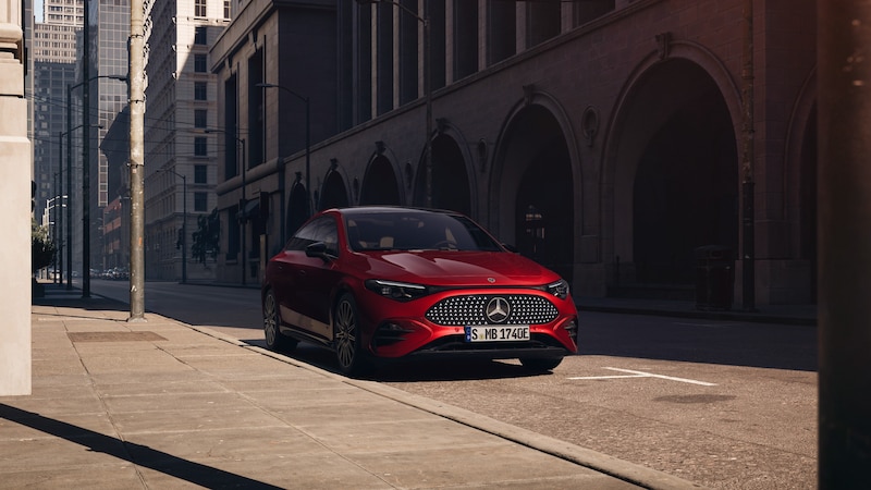 Ein roter Mercedes-Benz CLA Elektrisch fährt bei Sonnenlicht durch eine städtische Straße mit hohen Gebäuden und Arkaden.