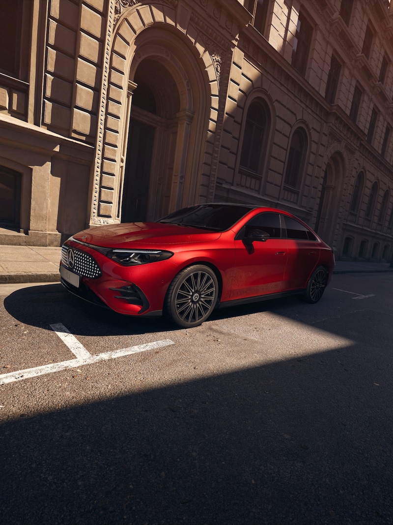 Ein roter Mercedes-Benz CLA Elektrisch steht in einer schattigen Stadtstraße, wobei Frontpartie, seitliche Silhouette und dunkle Räder sichtbar sind.
