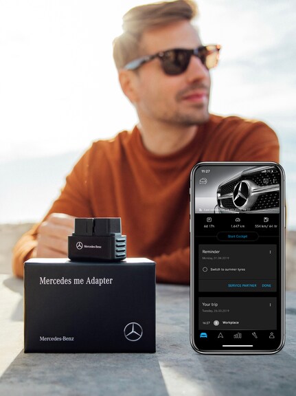 Accident detection with the Mercedes-Benz Adapter. | Mercedes-Benz A man wearing sunglasses sitting at a table with a Mercedes-Benz Adapter and a smartphone with the Mercedes-Benz Adapter app open in the foreground.
