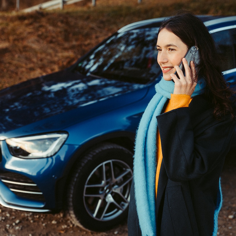 Roadside Assistance | Mercedes-Benz A woman standing in front of her Mercedes SUV, talking on a smartphone.