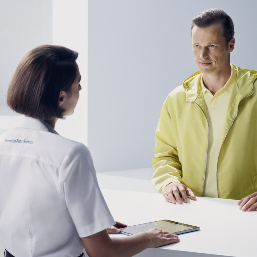 A customer leaning at the counter turns expectantly to face a Mercedes-Benz employee.