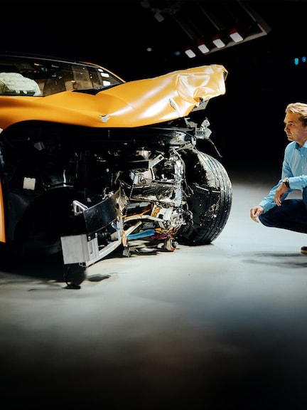 Crash test experts | Safety | Mercedes-Benz Crash test expert examing front-crashed Mercedes-Benz car while crouching bedite it.