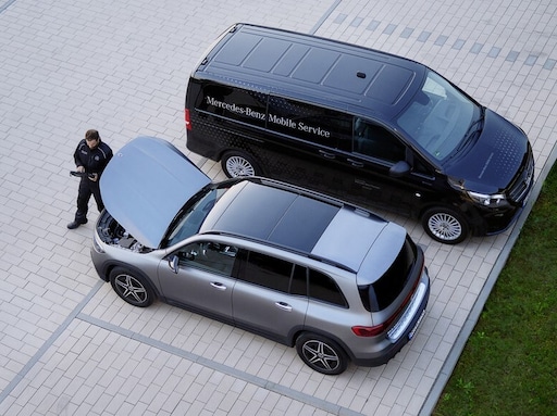 Un utilitaire léger Mercedes-Benz Mobile Service garé sur une allée pavée à côté d'un SUV argenté dont le capot moteur est ouvert, tandis qu'un technicien se tient à proximité, tenant une tablette de diagnostic.